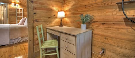 A wooden cabin room with a bed, a desk with a lamp, a chair, and a shelf with a plant.
