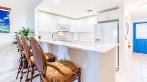 A modern kitchen with white cabinetry, a built-in microwave, and a refrigerator. There is a dining area with wicker chairs and a table. - Oceanfront right next to RumPoint, tranquility,pool,gym,tennis court and sunsets (Rum Point)
