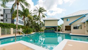 A swimming pool with a clear blue water, surrounded by palm trees and modern buildings. - Oceanfront right next to RumPoint, tranquility,pool,gym,tennis court and sunsets (Rum Point)