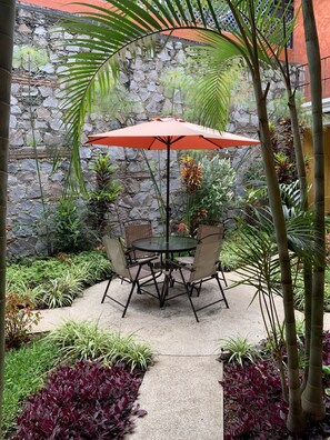 A patio with a stone wall, a round table with chairs, and an orange umbrella. - Casa El Conquistador (Antigua Guatemala)