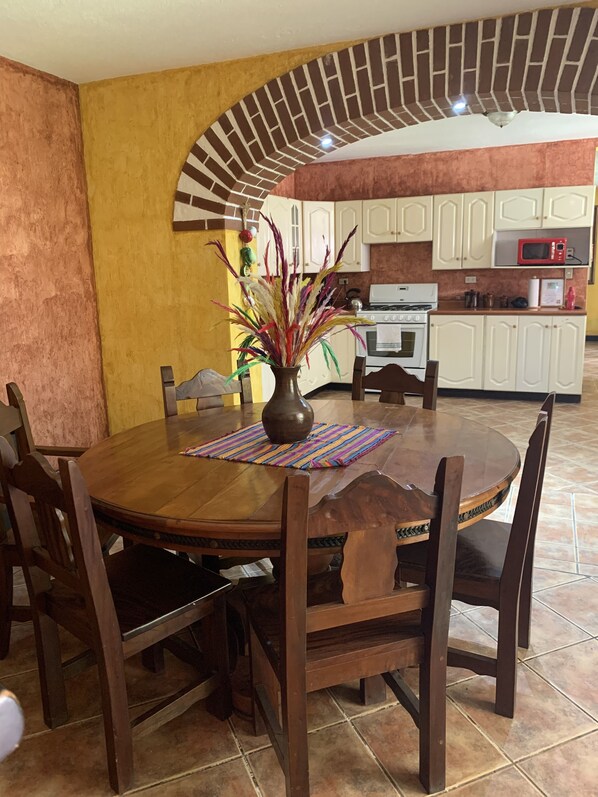 A dining area with a round wooden table, chairs, and a vase with decorative plants. - Casa El Conquistador (Antigua Guatemala)