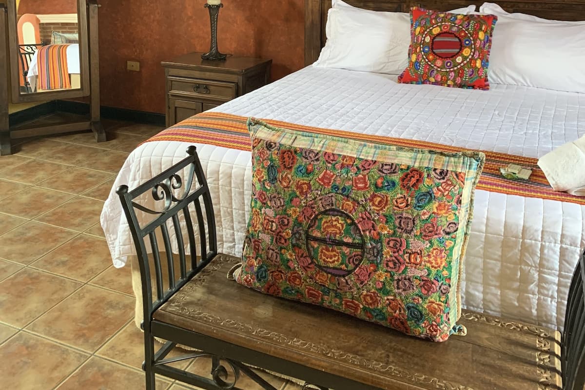 Ein Schlafzimmer mit Bett, Nachttisch, Lampe, Stuhl und einem farbenfrohen Bild an der Wand.