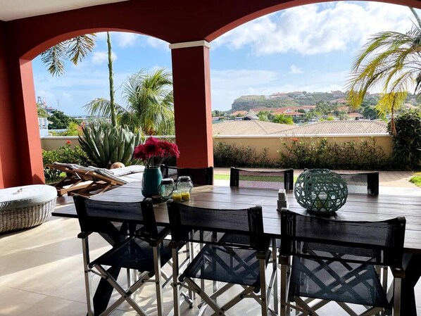 An outdoor patio with a table and chairs, surrounded by plants and a view of a hilly landscape.