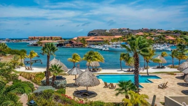 Une station balnéaire dotée d’une piscine, de palmiers et d’un port de plaisance où sont amarrés des bateaux.