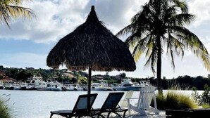 Une plage avec des chaises longues et un parasol en chaume, un palmier et des bateaux en arrière-plan.