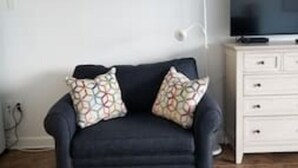 Een donkere bank met gekleurde kussens, een witte lamp en een televisie op een wit kastje in een kamer met betegelde vloer.