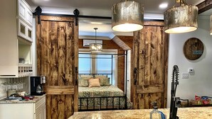 Una cocina con encimera de granito, cafetera y fregadero. Se ve una cama con colcha estampada y almohadas al fondo.