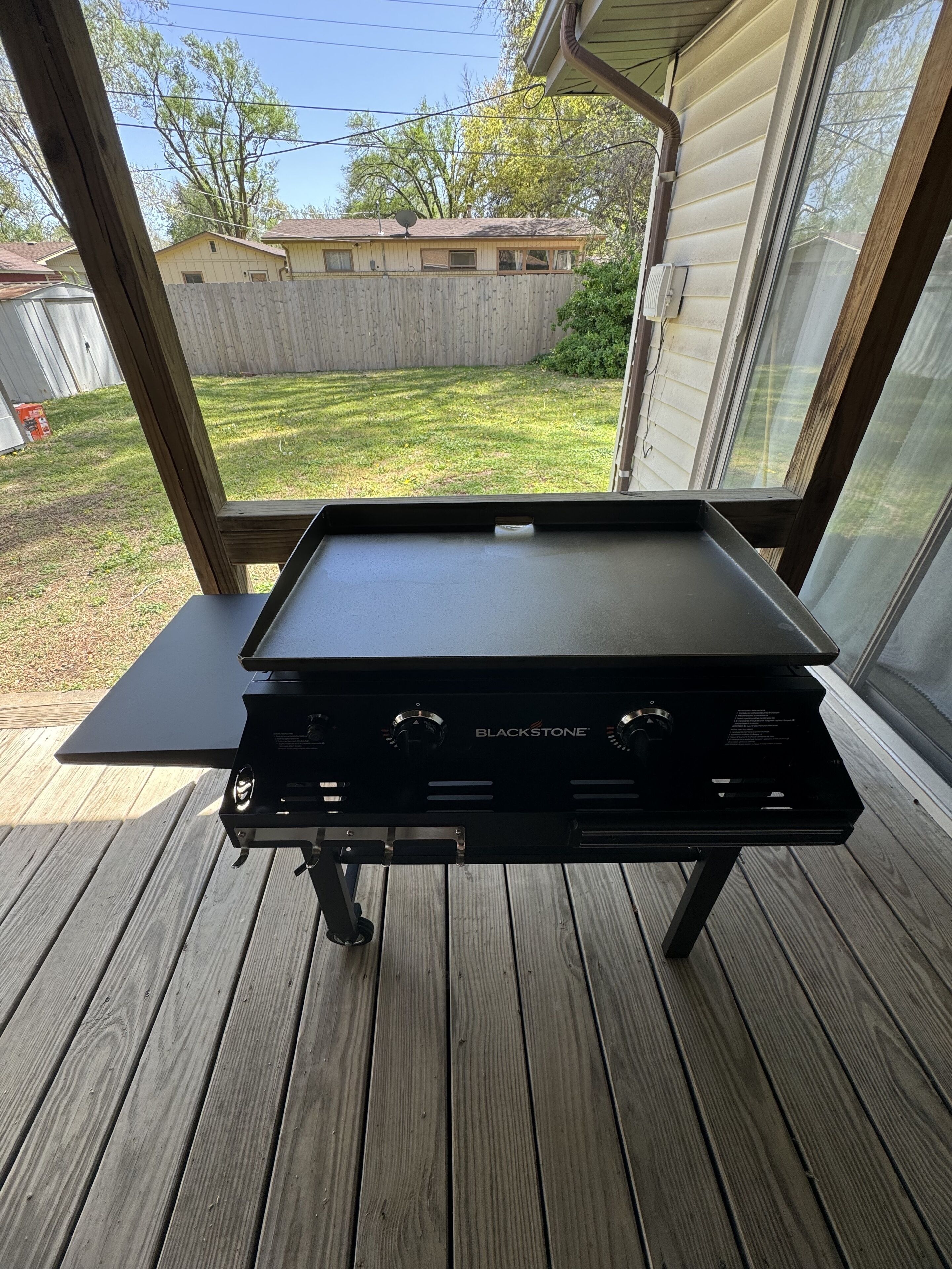 Un barbecue Blackstone sur une terrasse en bois, avec vue sur un jardin et une maison.