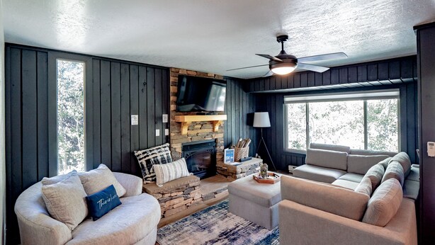 A cozy living room with a stone fireplace, a ceiling fan, and a large window providing natural light.