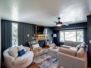A cozy living room with a stone fireplace, a ceiling fan, and a large window providing natural light.