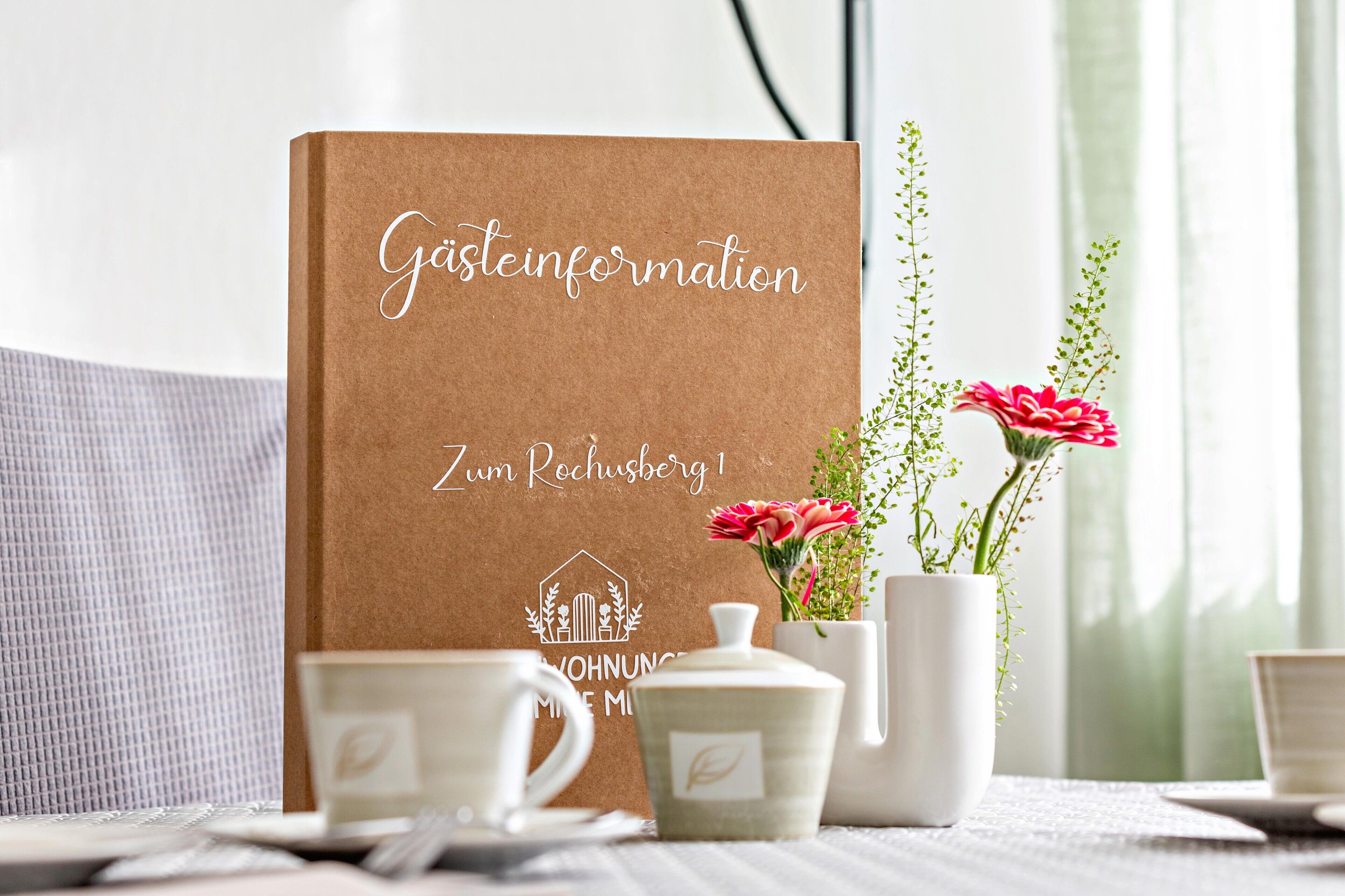Ein braunes Buch mit dem Titel 'Gästeneinladung Zum Rochusberg 1' und einem Logo, platziert auf einem Tisch zusammen mit einer weißen Tasse, einem weißen Teekessel und einer Blumenvase.