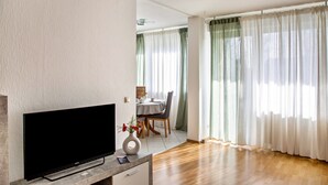 A living room with a flat-screen TV, a dining area with a table and chairs, and large windows with sheer curtains.