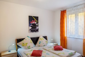 Una camera da letto con un letto, cuscini, un comodino, una lampada, una finestra con tende e un quadro appeso al muro.