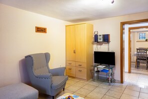 Una sala de estar con un sillón a cuadros, un armario de madera, un mueble de televisión de vidrio y un área de comedor visible a través de un umbral abierto.