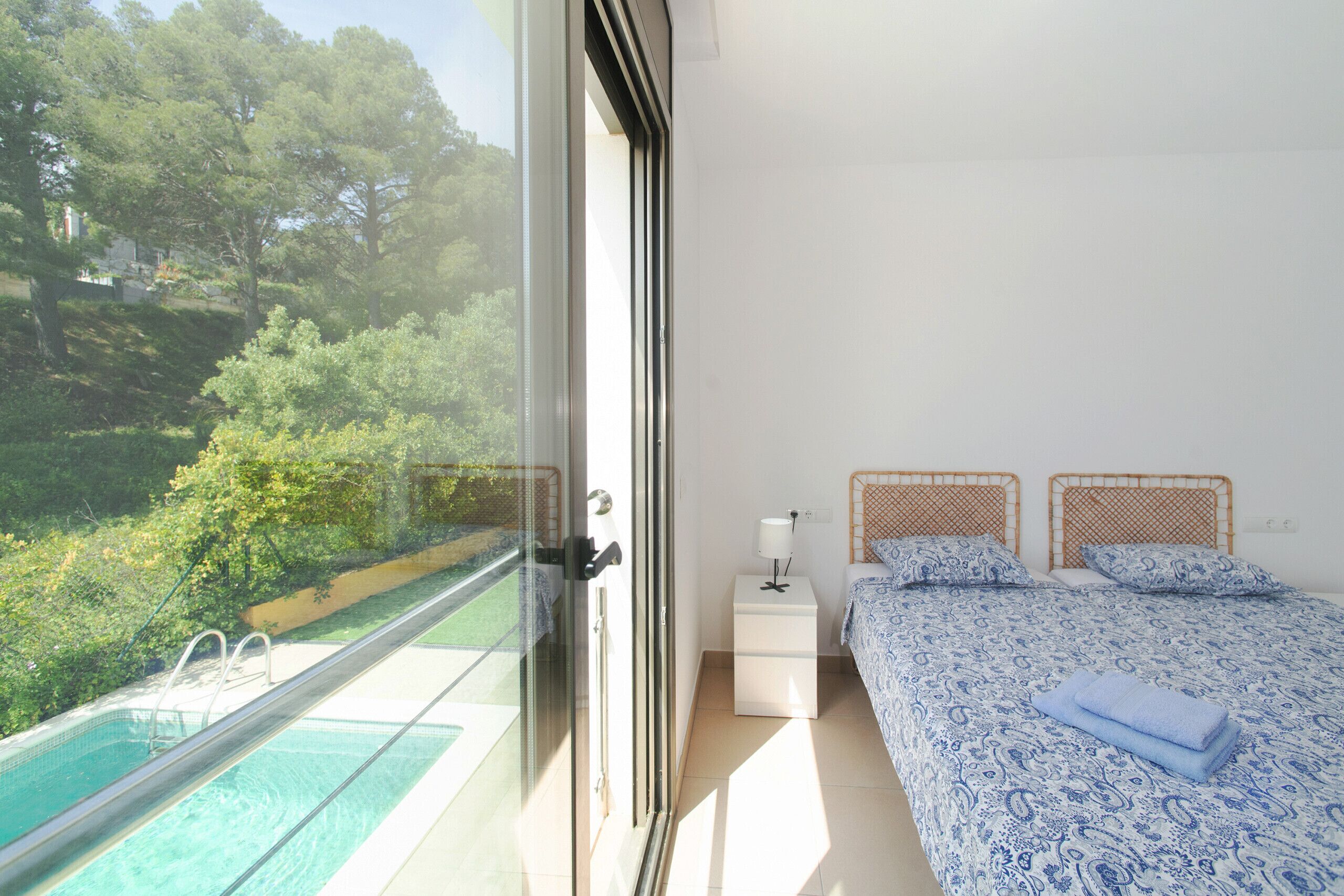 A bedroom with a view of a swimming pool, a bed with patterned bedding, a bedside table with a lamp, and a window with a view of greenery.