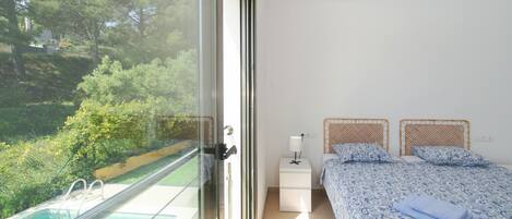 A bedroom with a view of a swimming pool, a bed with patterned bedding, a bedside table with a lamp, and a window with a view of greenery.