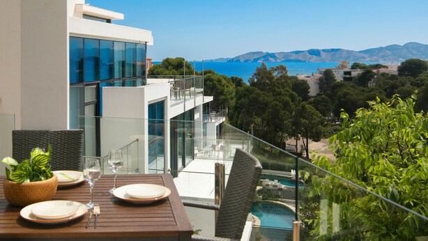 Un área de comedor exterior moderna con vista al mar y a las montañas, barandilla de cristal y piscina.