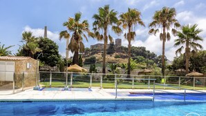 Une piscine à l’eau cristalline, entourée de palmiers, avec en toile de fond une colline surmontée d’une structure ressemblant à un château.