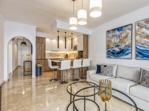 A modern living room with a sofa, a glass coffee table, and a kitchen area with bar stools. - Rio | sea views - terrace - pool (Fuengirola)