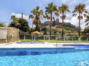 A swimming pool with clear blue water, surrounded by palm trees and a view of a hill with a castle-like structure in the background. - Rio | sea views - terrace - pool (Fuengirola)