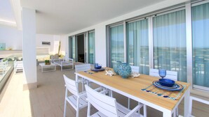 A modern outdoor dining area with a wooden table and white chairs, large windows with curtains, and a view of the cityscape. - 2202 Modern Apartment with Panoramic Sea Views (Cancelada Estepona)