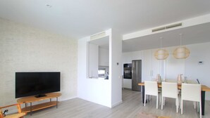 A modern living room with a flat-screen TV on a wooden stand, a dining area with white chairs and a wooden table, and a kitchen in the background. - 2202 Modern Apartment with Panoramic Sea Views (Cancelada Estepona)