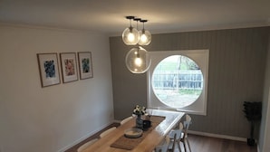 Un comedor con mesa de madera, sillas blancas, un ventanal redondo con vista a un patio, y tres obras de arte enmarcadas en la pared.