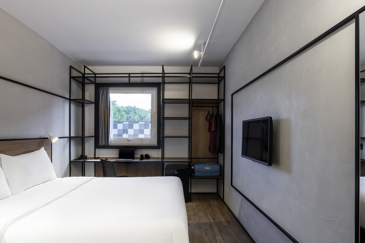 A modern hotel room with a bed, a desk with a computer, a TV, and a wardrobe.