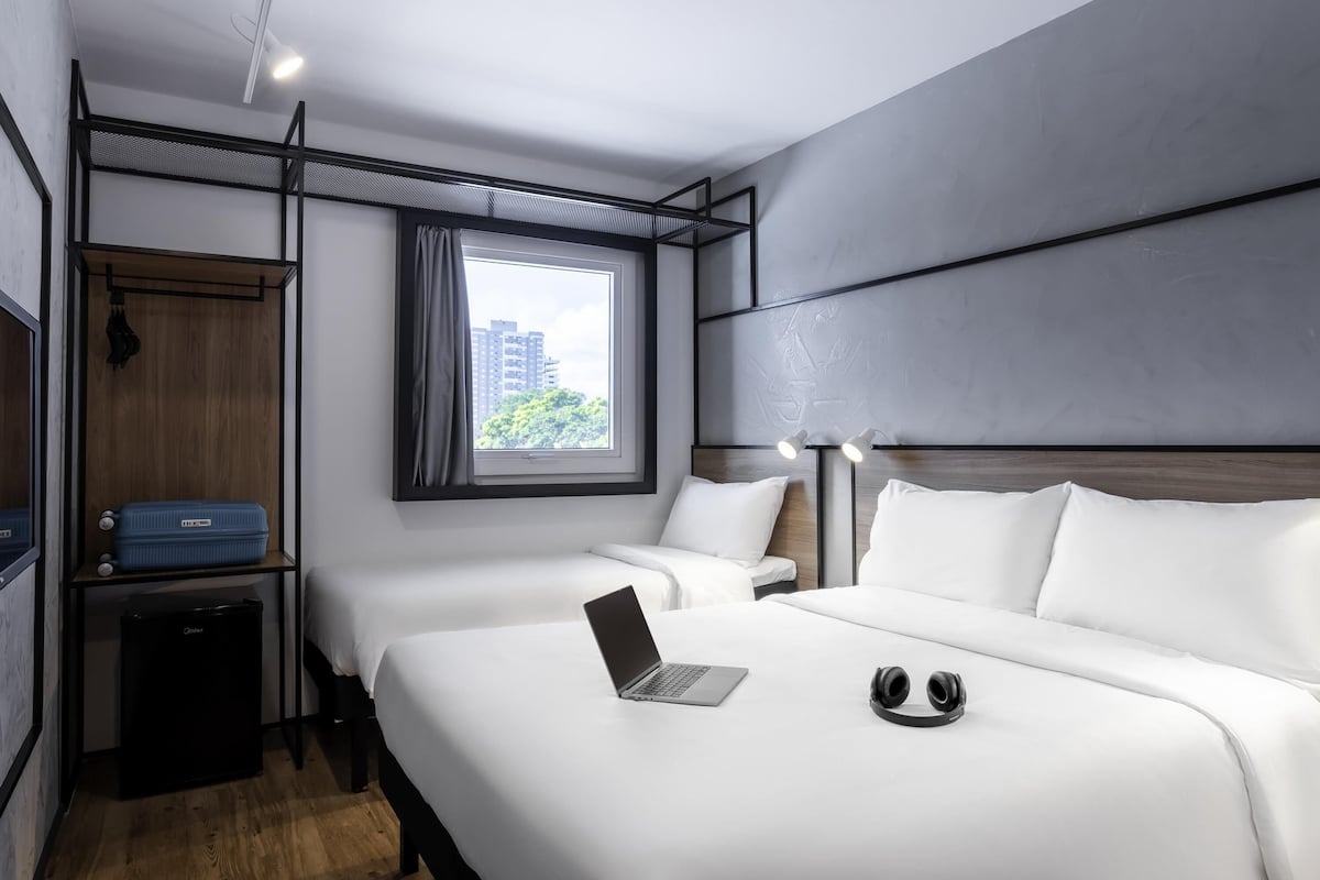 A modern hotel room with two beds, a desk, a laptop, and a view of buildings through a window.
