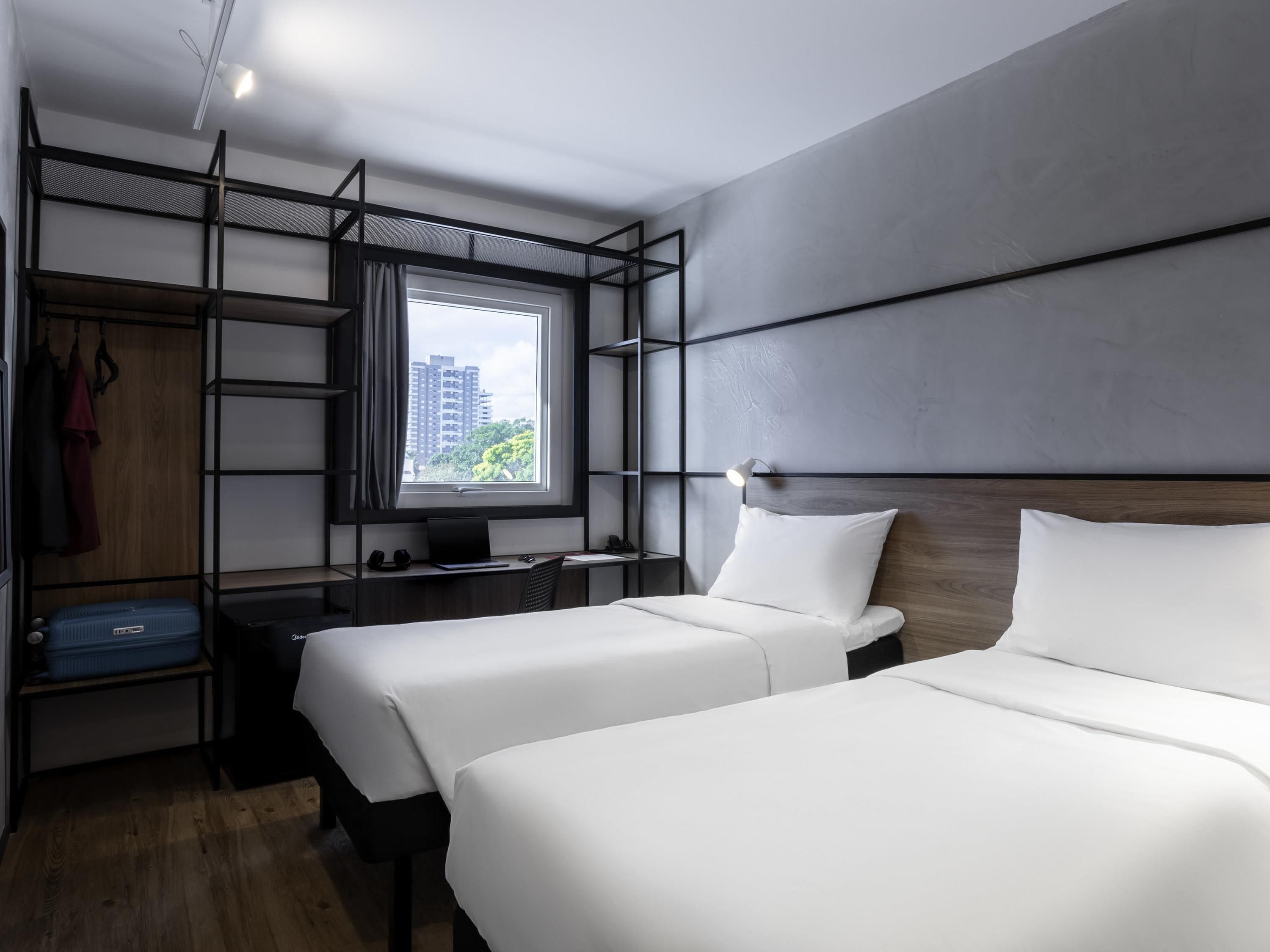 Ein modernes Hotelzimmer mit zwei Betten, einem Schreibtisch, einem Kleiderschrank und einem großen Fenster mit Blick auf die Stadt.