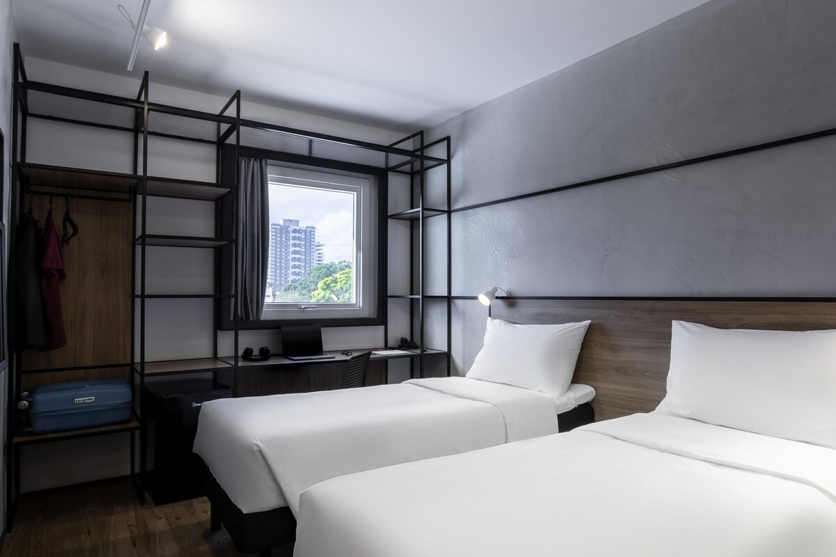 A modern hotel room with two beds, a desk, a wardrobe, and a large window offering a city view.