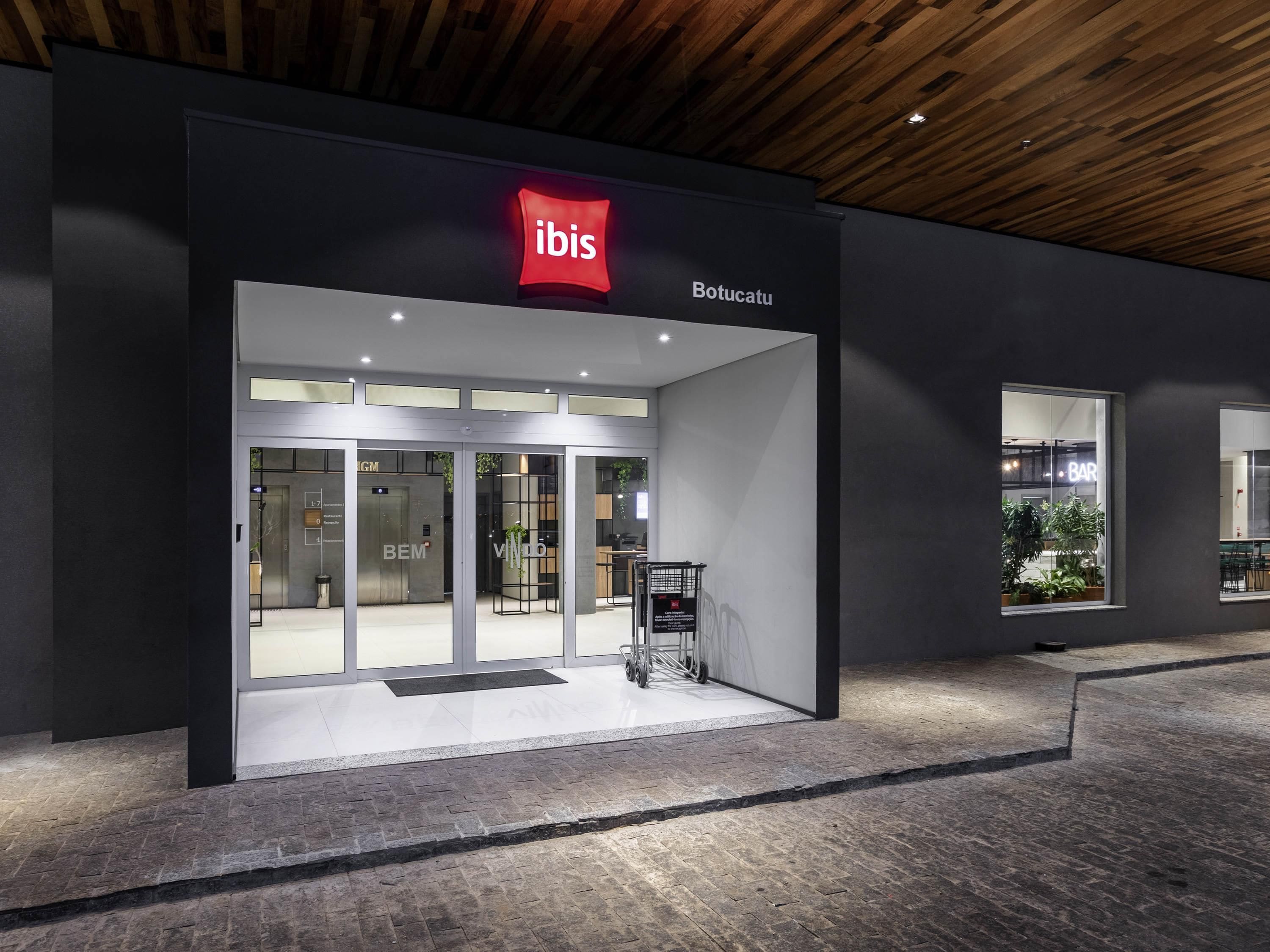 A modern hotel entrance with a prominent 'ibis' logo, a glass door entrance, and a visible elevator.