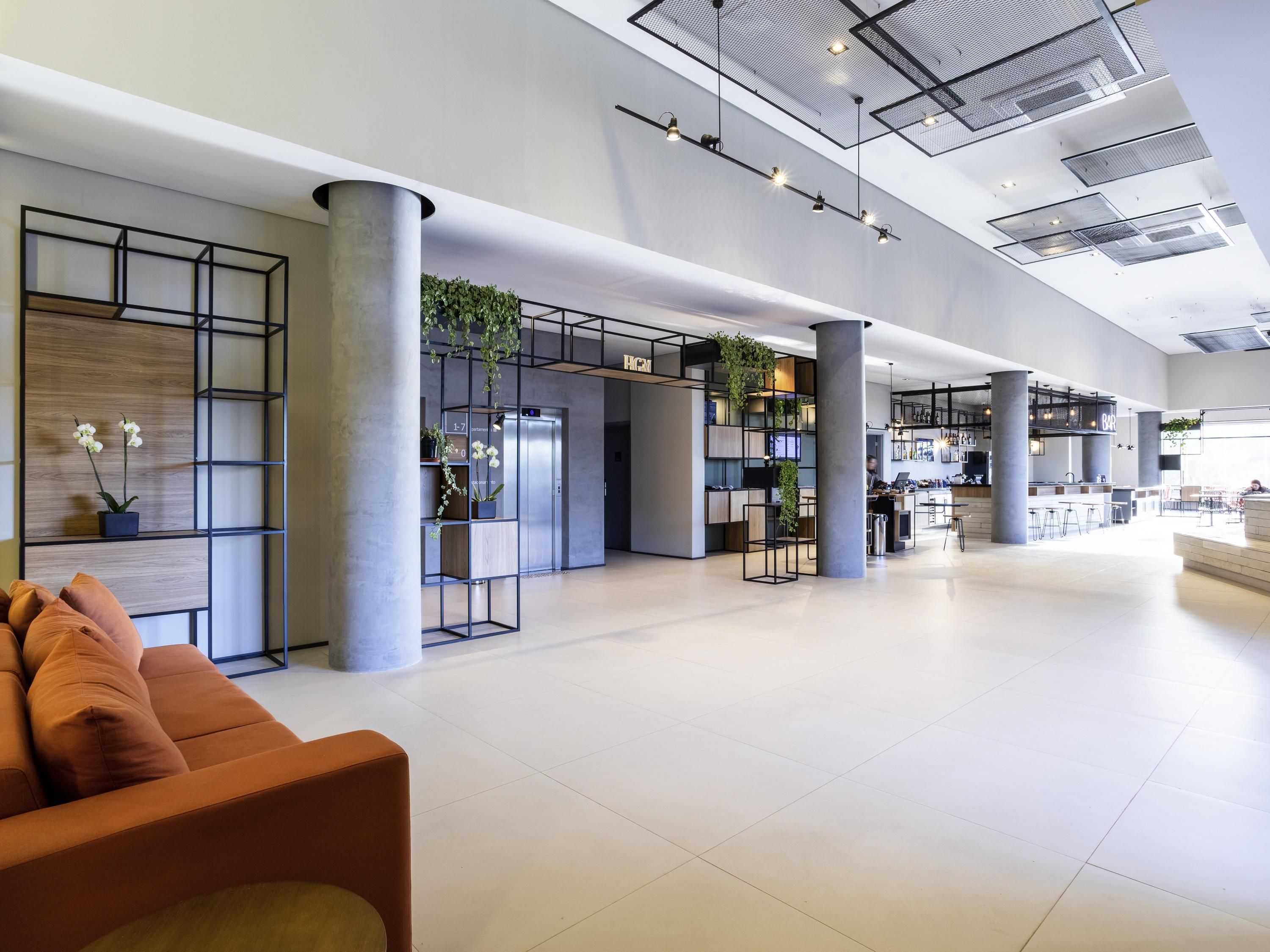 A spacious lobby with a modern design, featuring a seating area with orange cushions, a reception desk, and a bar area.