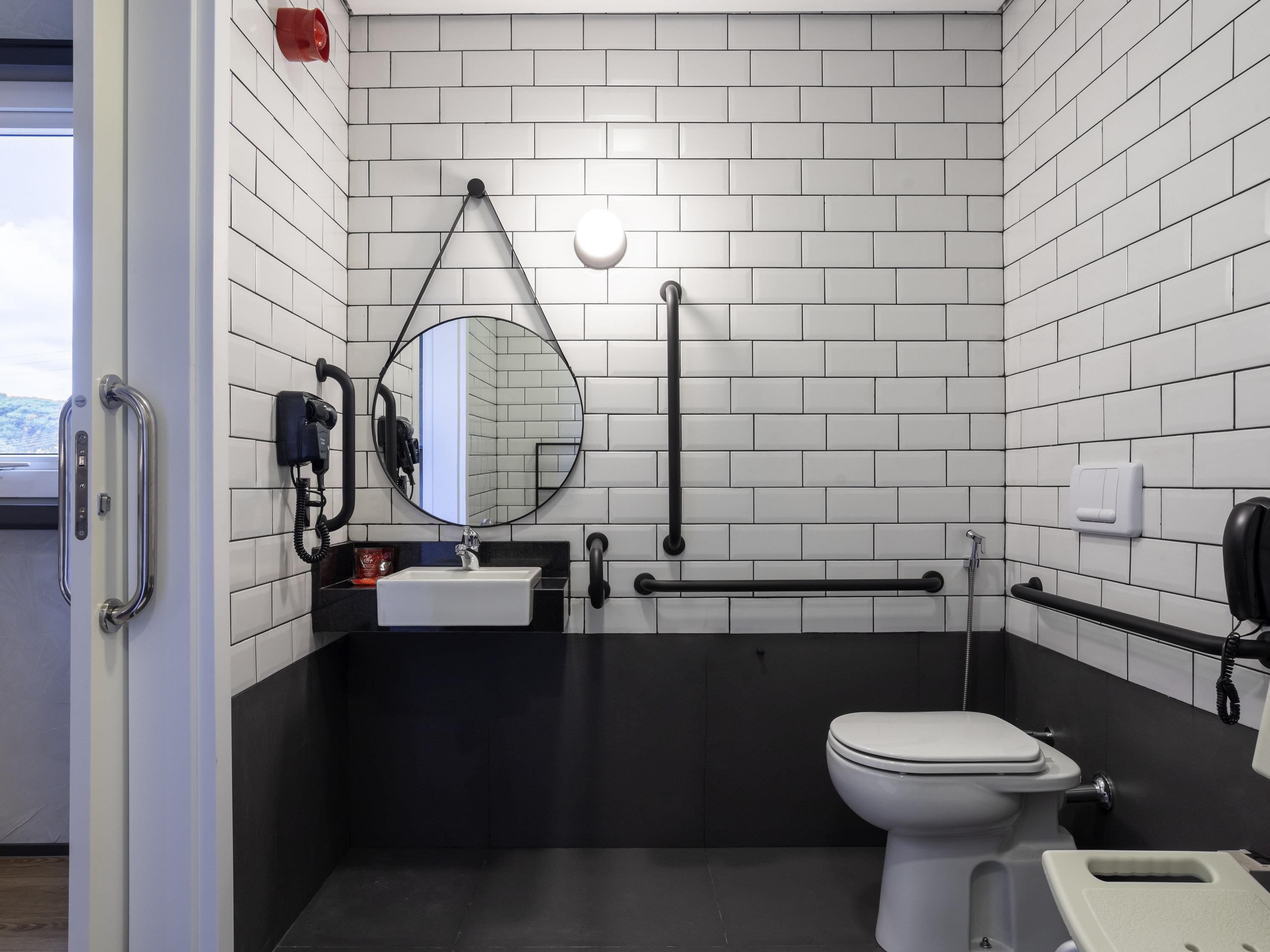 Et moderne badeværelse med hvide fliser, et hvidt toilet, en vask med spejl og et gelænder.
