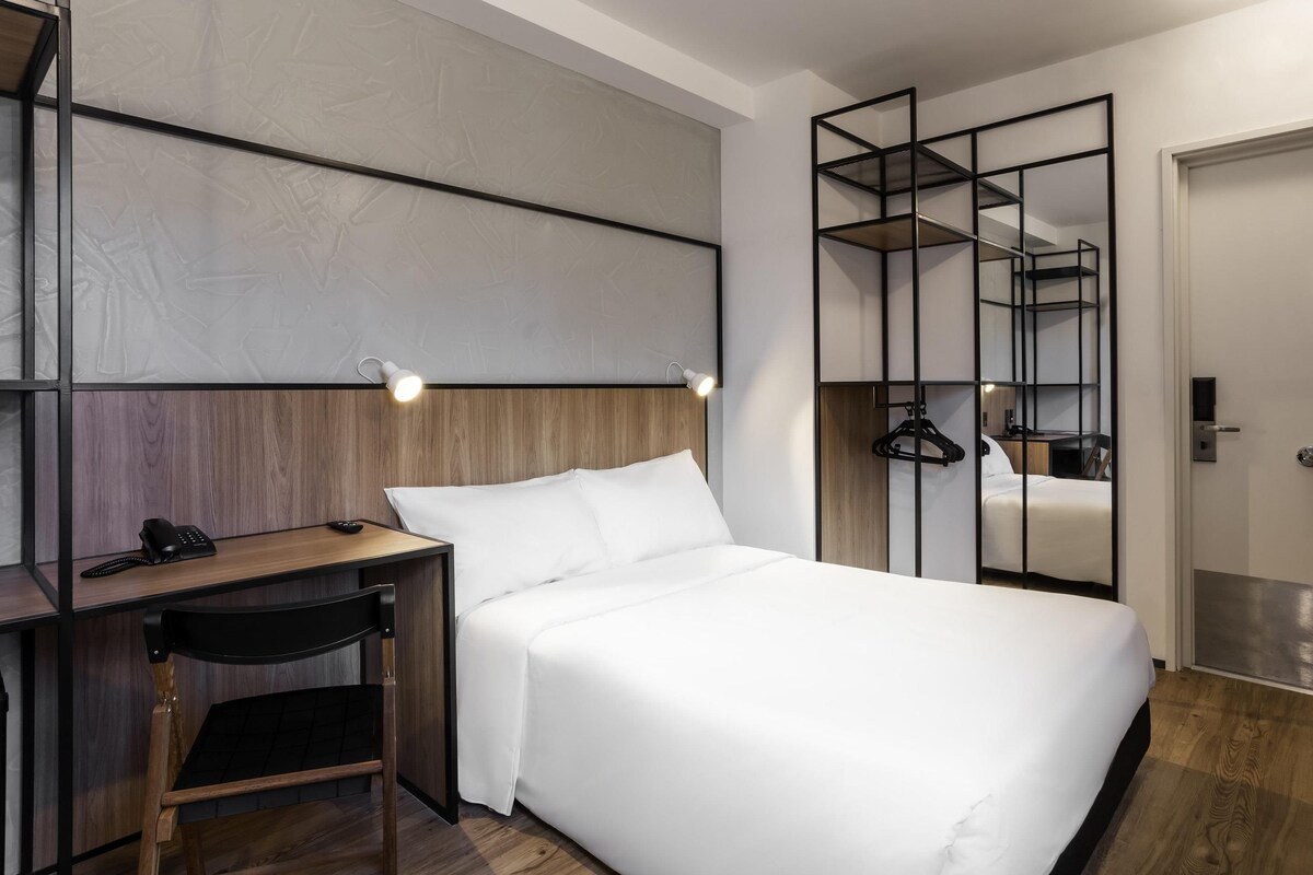 A hotel room with a bed, a desk, a chair, a phone, and a mirror.