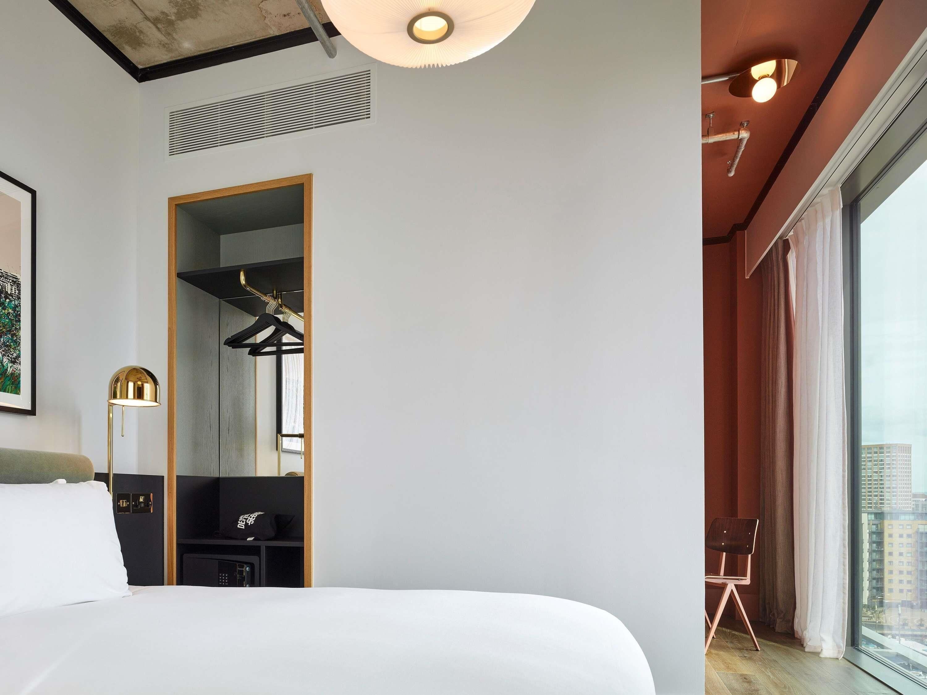 Ein Hotelzimmer mit einem großen Bett, einem Schrank, einem Spiegel, einem Schreibtisch und Blick auf die Stadt durch das Fenster.