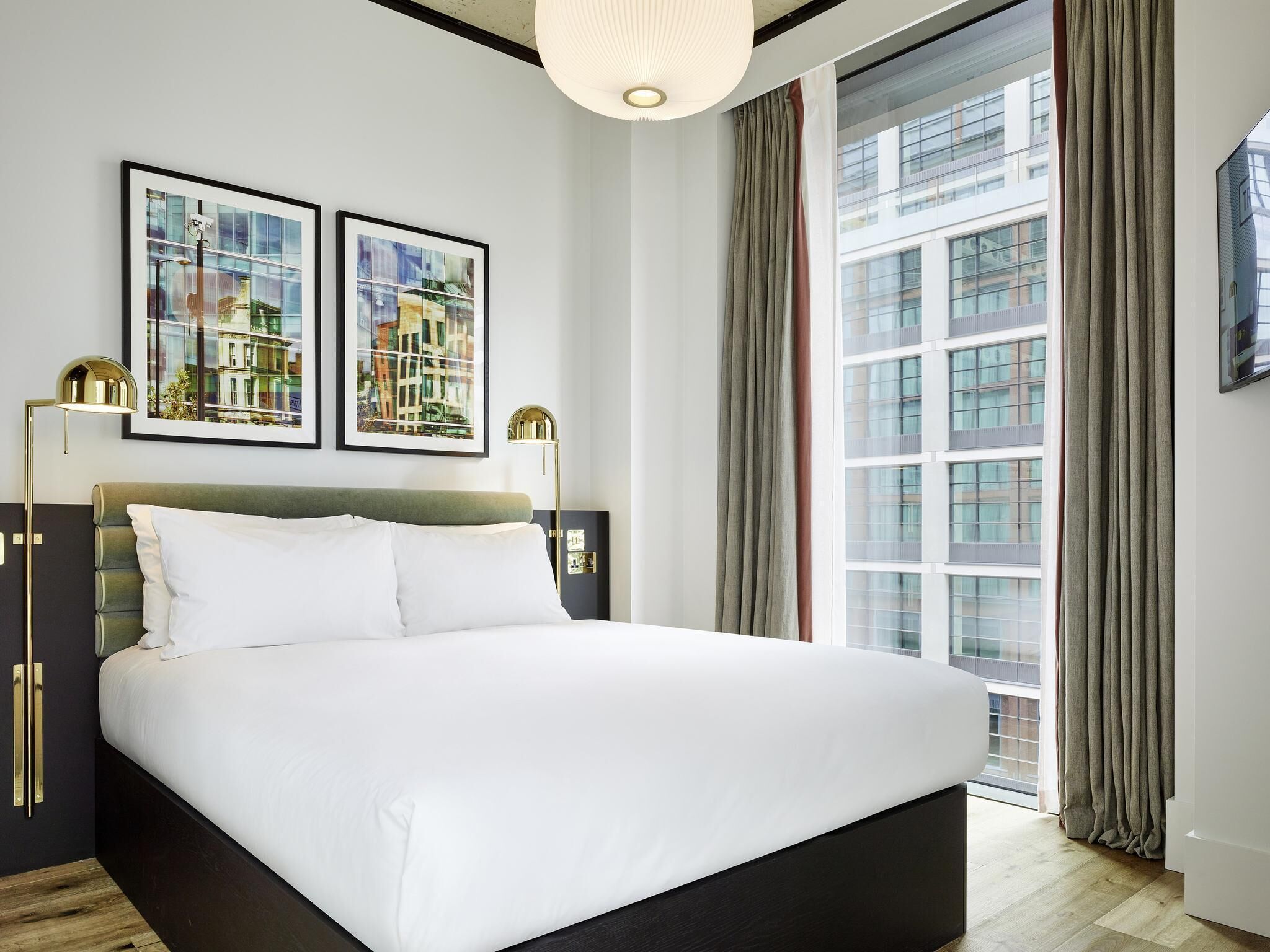 A modern hotel room with a large bed, two framed cityscape pictures, a floor lamp, and a view of a building through the window.
