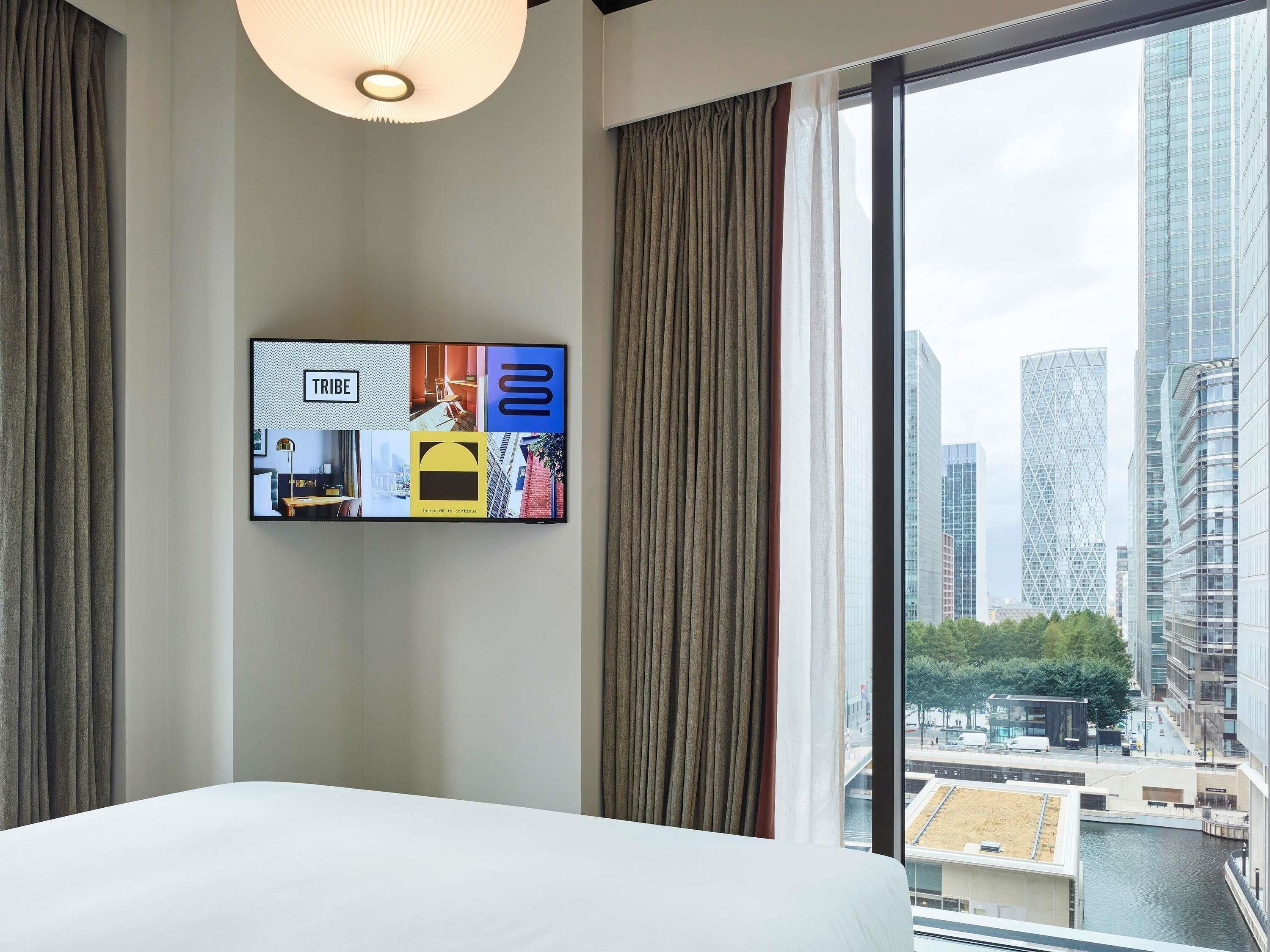 Ein Hotelzimmer mit einem großen Bett, einem an der Wand befestigten Fernseher, der verschiedene Bilder zeigt, und Blick auf eine Stadtlandschaft durch ein großes Fenster.