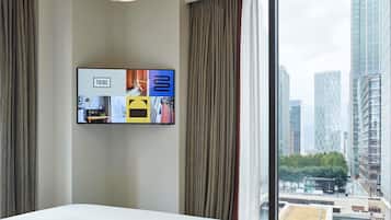 Habitación de hotel con una cama grande, un televisor montado en la pared mostrando varias imágenes y vistas a un paisaje urbano a través de un ventanal amplio.
