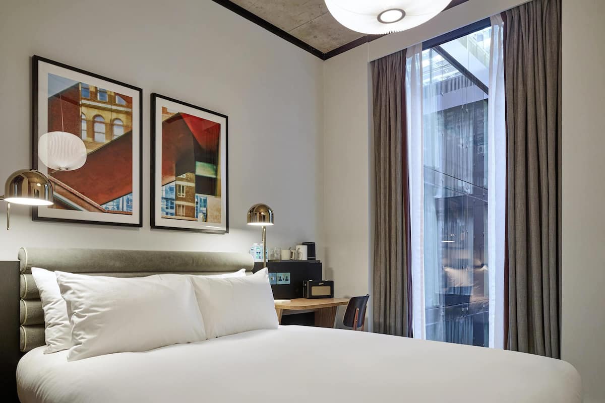 A hotel room with a large bed, two framed artworks, a desk with a radio, and a view of a building through the window.