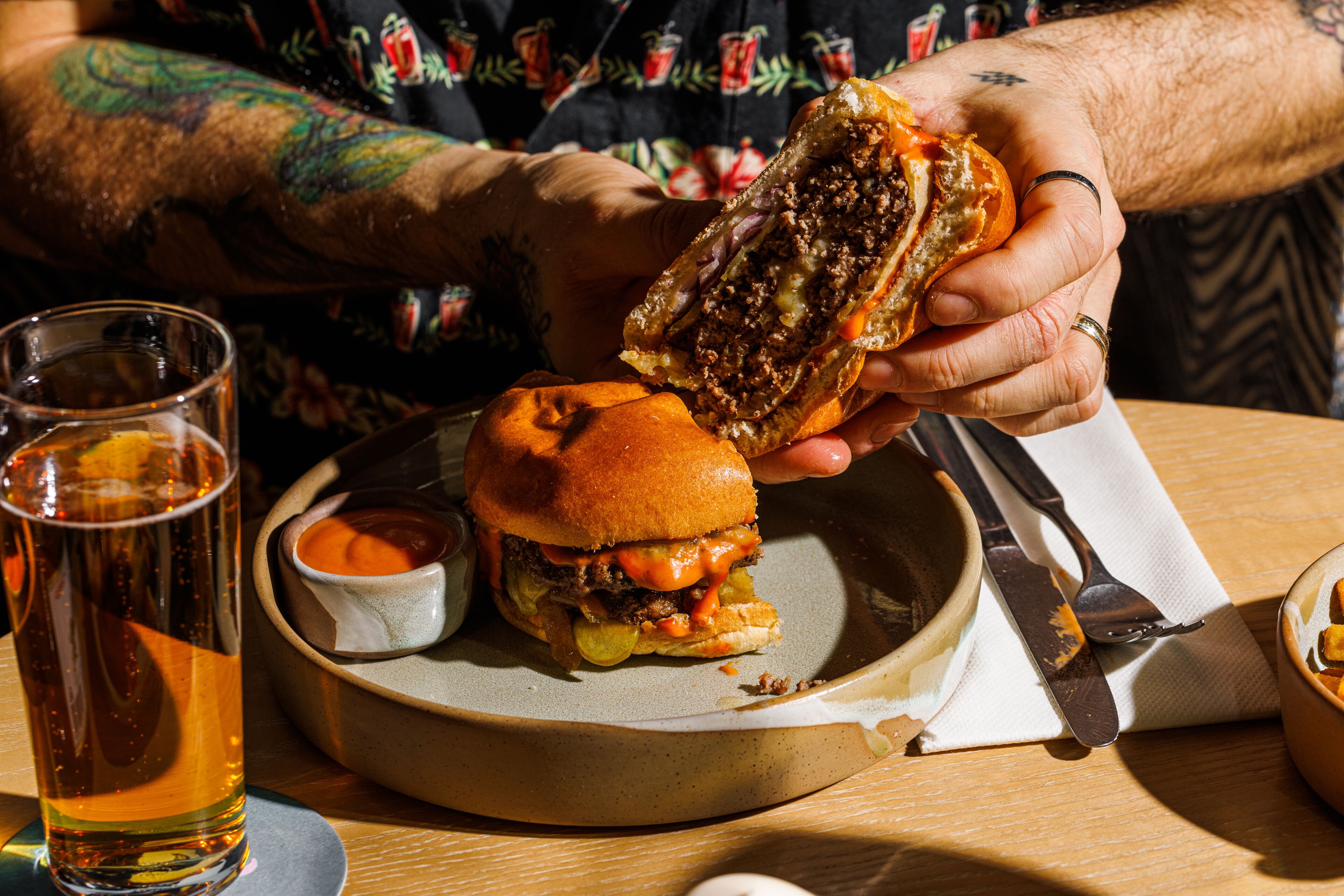 A person holding a sandwich with a bun, a glass of beer, and a small bowl of sauce on a plate.