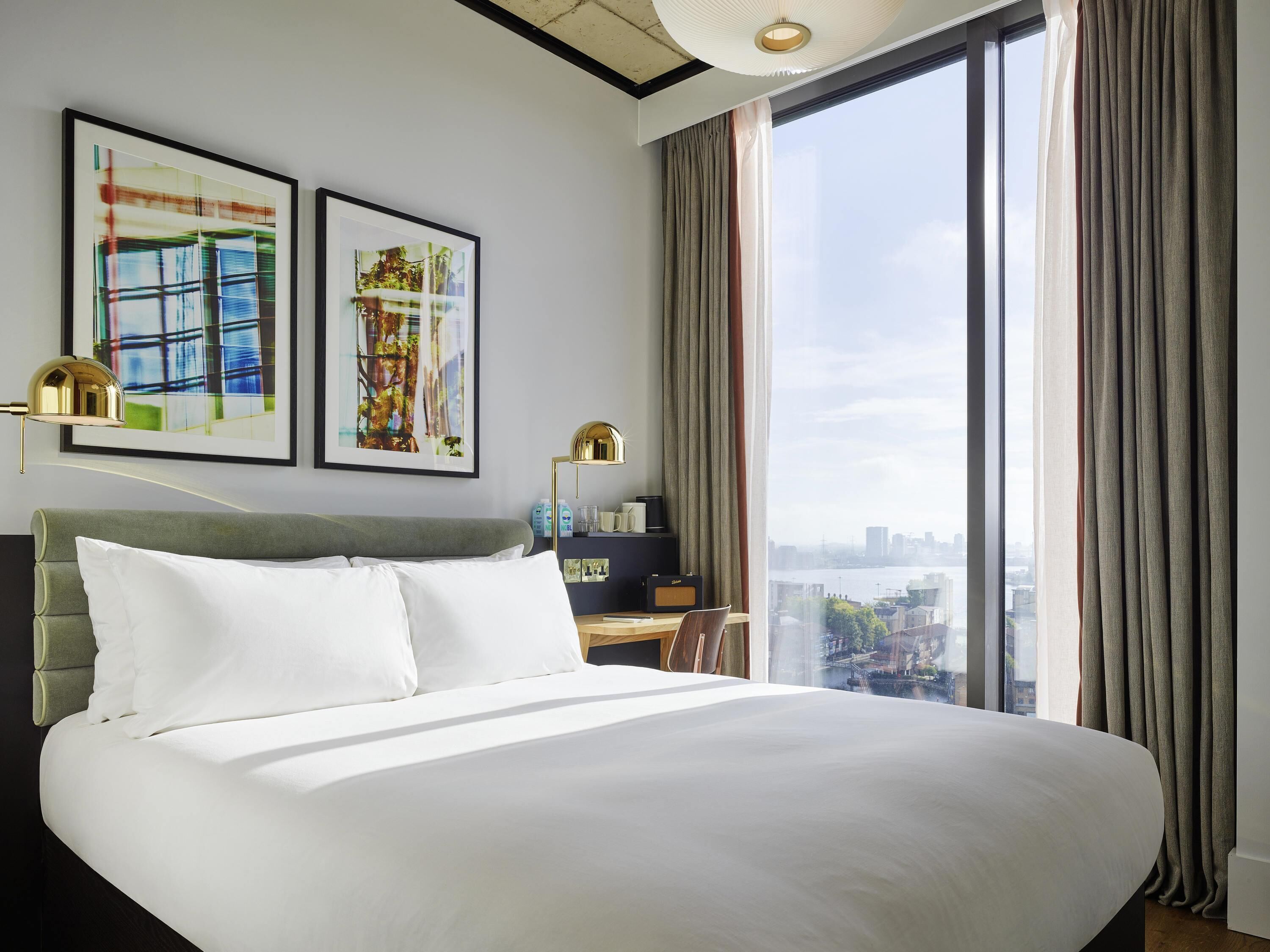 A hotel room with a large bed, two framed artworks, a desk with a lamp, and a view of a cityscape through large windows.