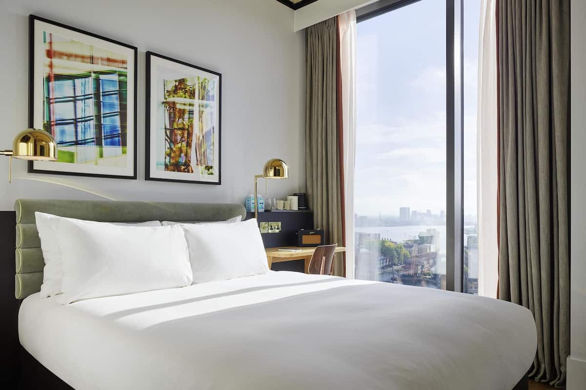 A hotel room with a large bed, two framed artworks, a desk with a lamp, and a view of a cityscape through large windows.