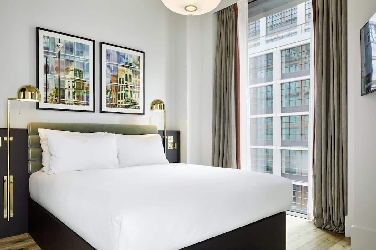 A modern hotel room with a large bed, two framed pictures on the wall, a floor lamp, and a view of a building through the window.