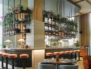 A bar area with multiple seating options, including stools and round tables, and a variety of plants and bottles on shelves. - Tribe London Canary Wharf (London)