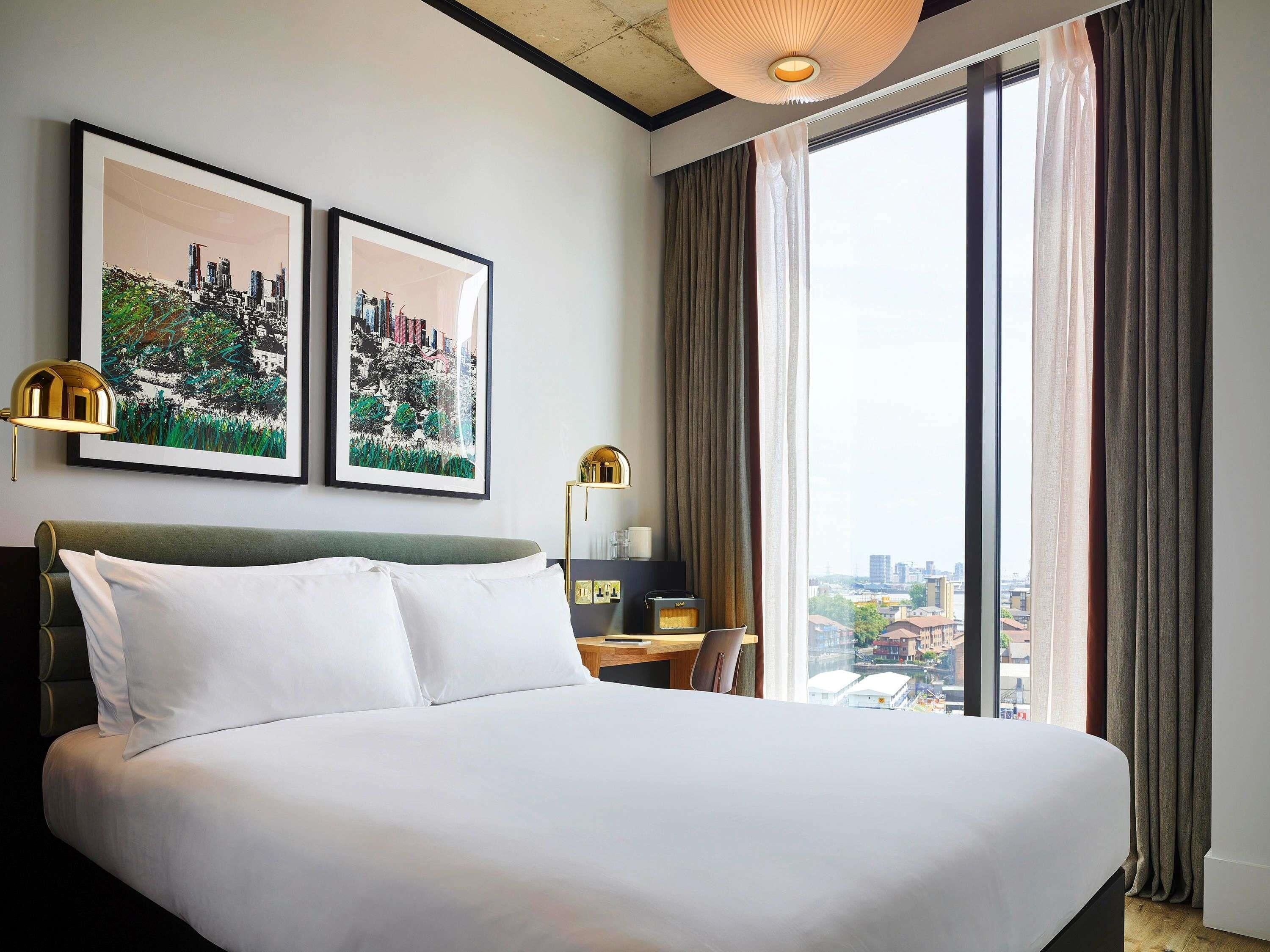A hotel room with a large bed, two framed cityscape pictures, a desk with a lamp, and a view of the city through the window.
