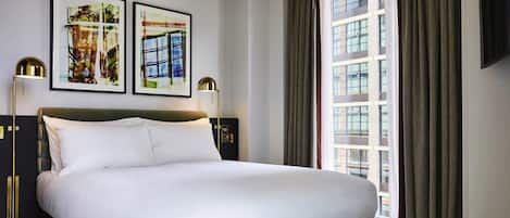 A modern hotel room with a large bed, two framed artworks, a floor lamp, and a view of the city through large windows.