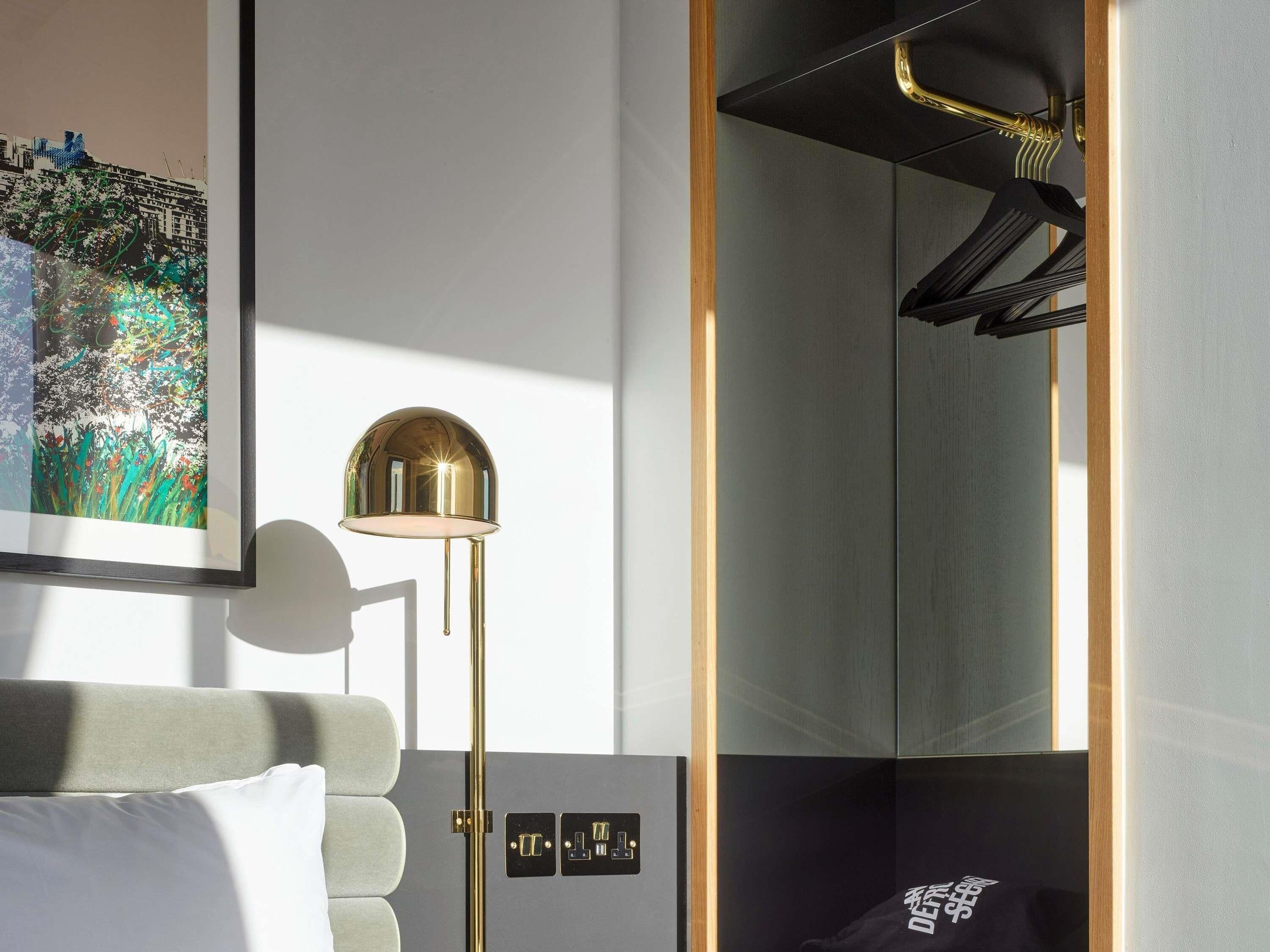 A modern hotel room with a bed, a floor lamp, a mirror, and a framed artwork on the wall.