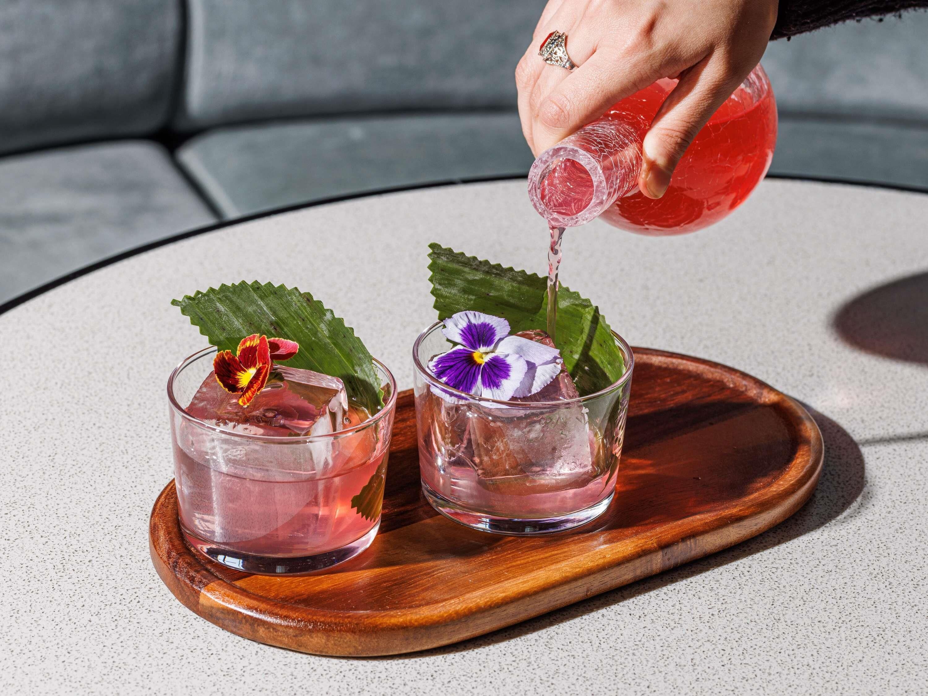 Eine Hand gießt ein Getränk in ein Glas, das mit einer Blüte verziert ist, auf einer Holztischplatte.