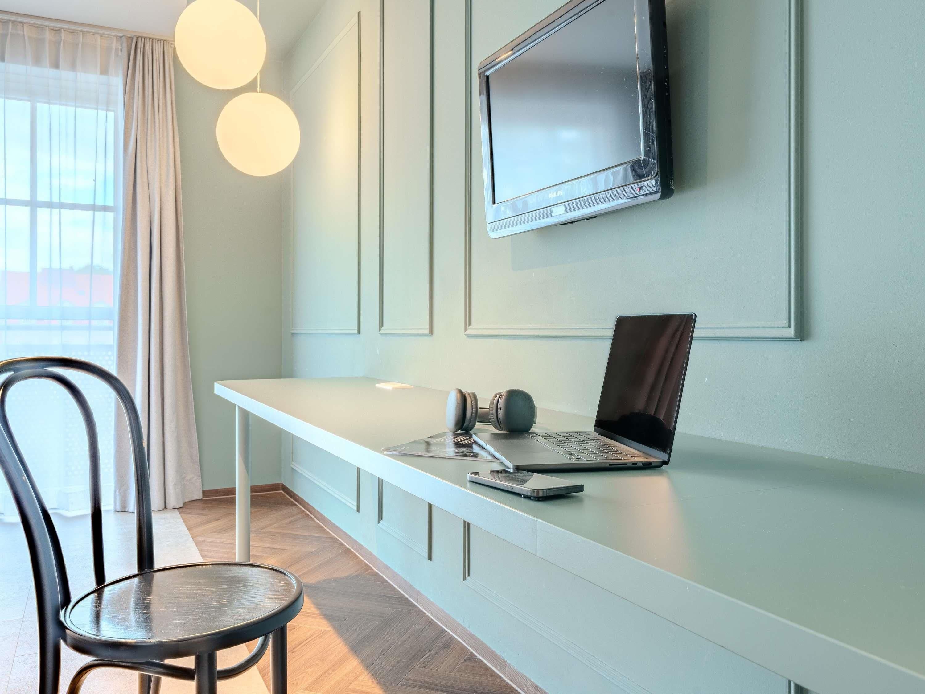 A modern office space with a white desk, a laptop, and headphones. A TV is mounted on the wall above the desk.